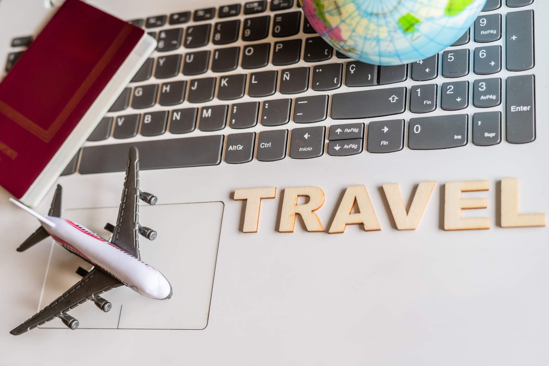 Travel planning concept featuring a small globe, passport, airplane model, and wooden letters spelling travel placed on a laptop keyboard, suggesting online booking and trip preparation