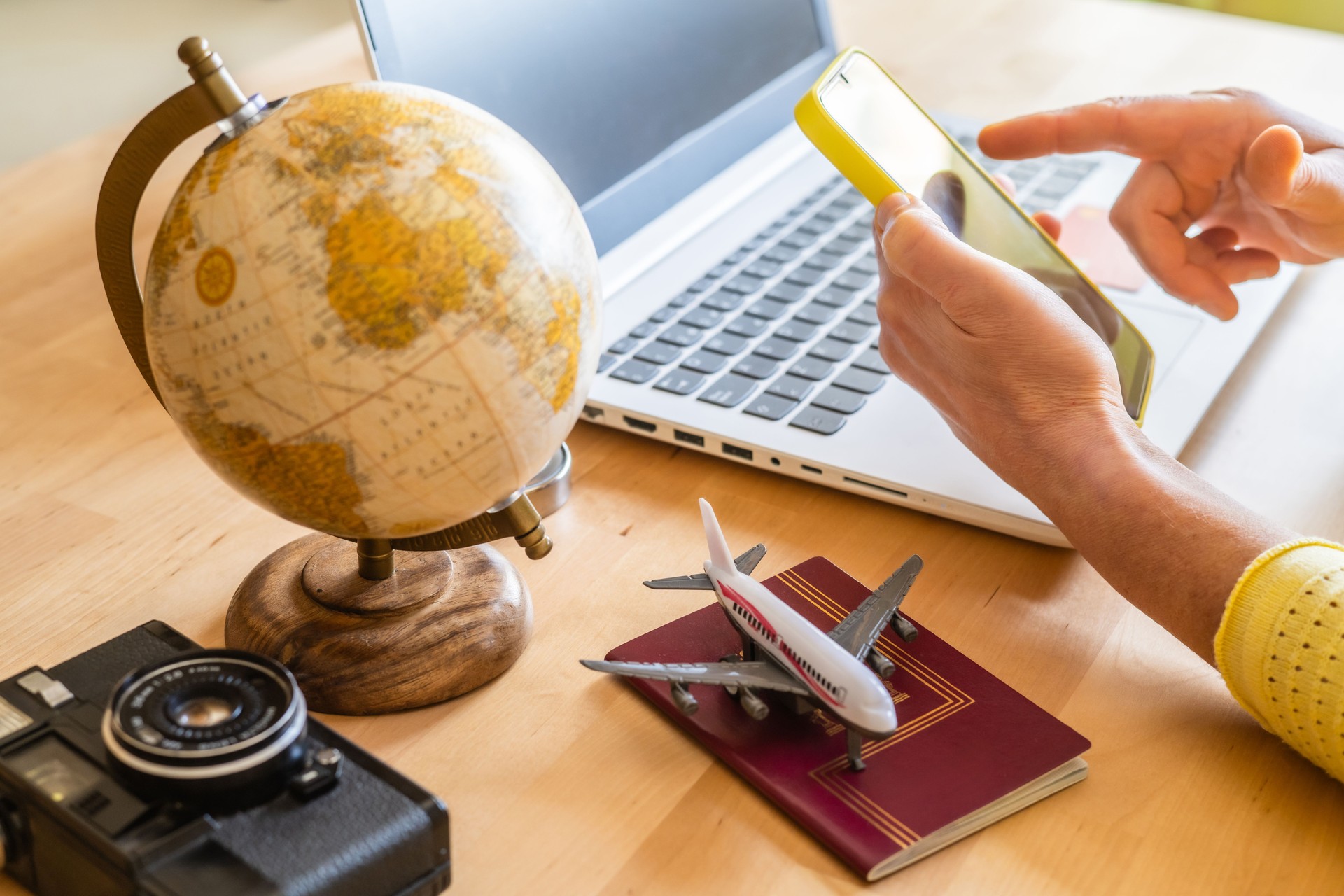 Travel agent using a smartphone and laptop for booking trips, surrounded by a globe, passport, toy airplane, and vintage camera on the desk, embodies modern travel planning